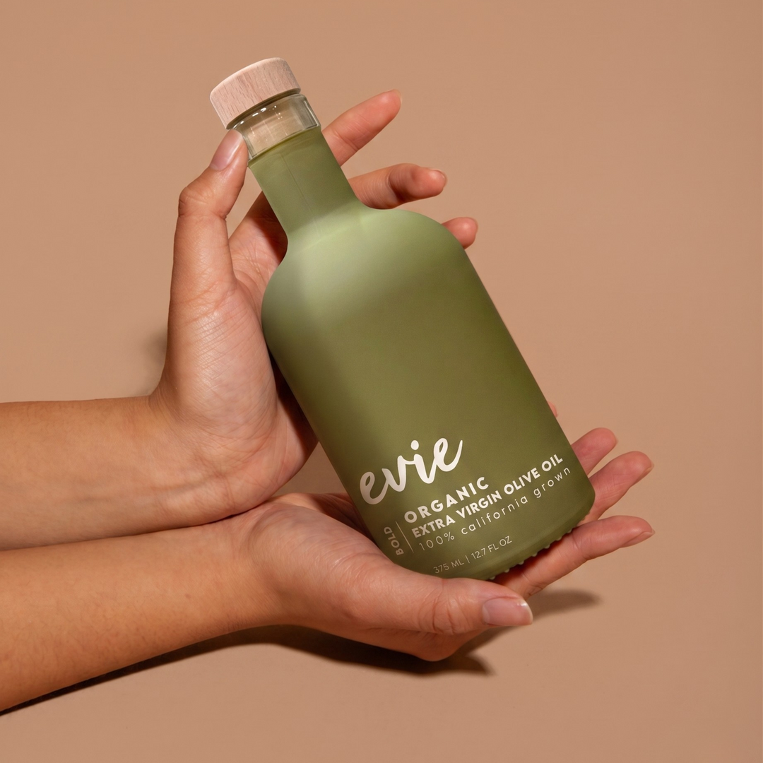 Person holding a green bottle labeled 'evie bold organic olive oil against a beige background