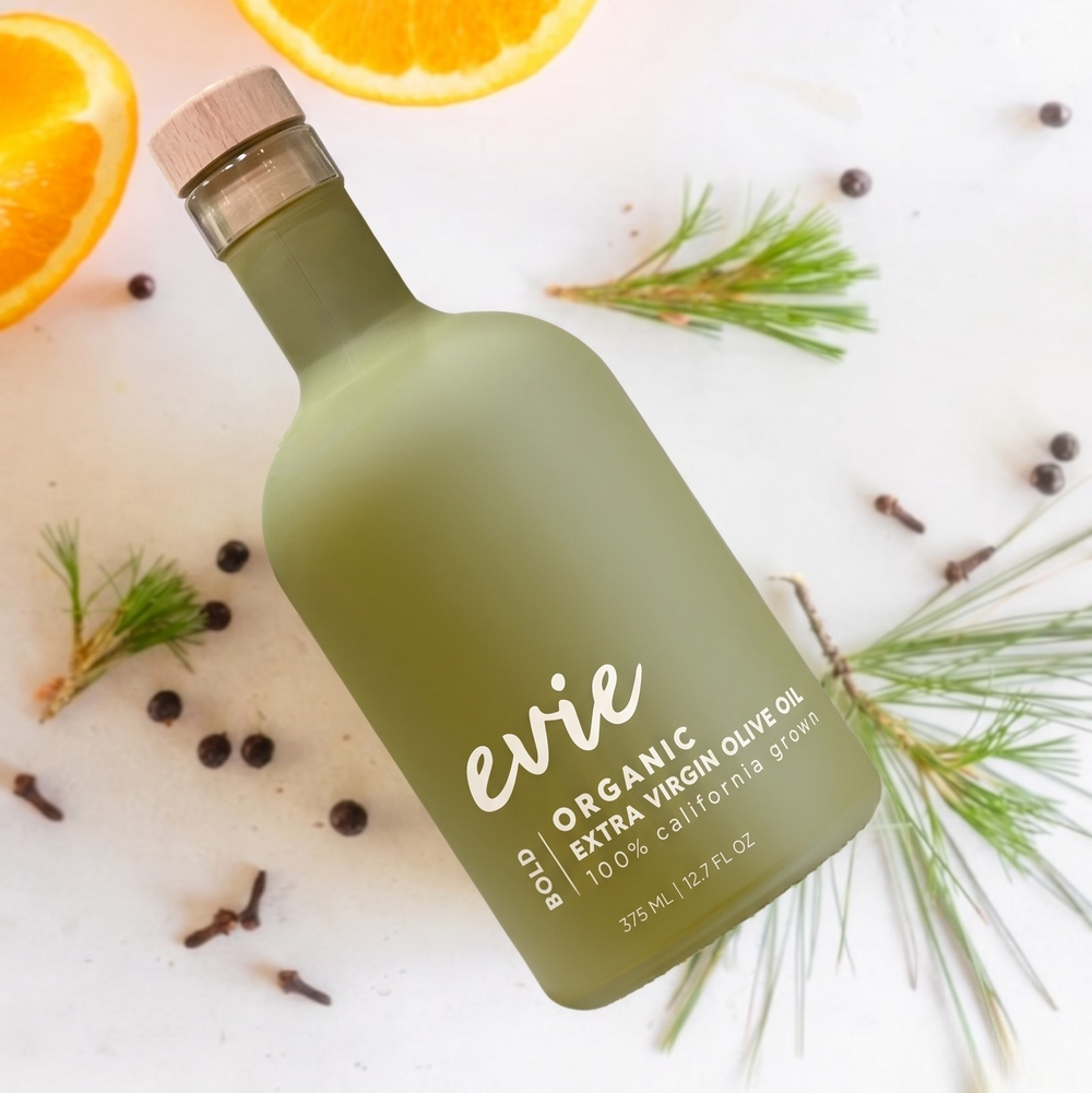 Green bottle labeled 'Evie Bold Organic Olive Oil with oranges and spices on a white surface