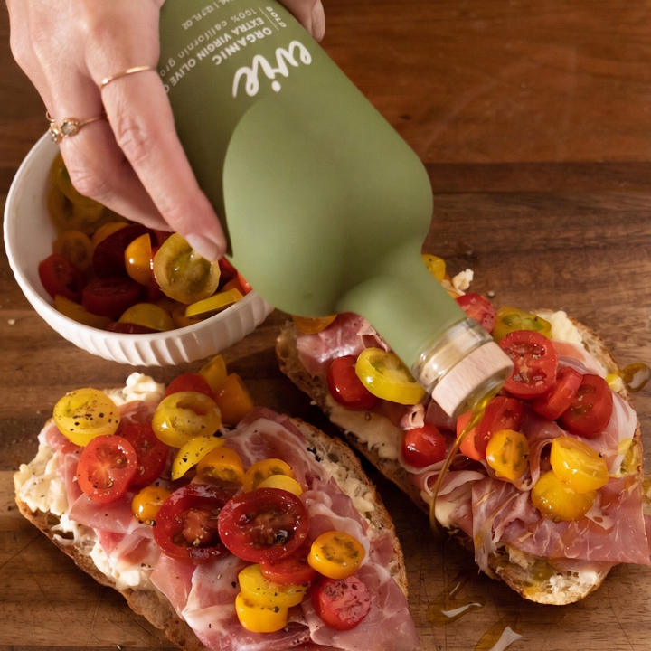 Person adding cherry tomatoes to a sandwich on a wooden surface with a bottle of evie organic olive oil.