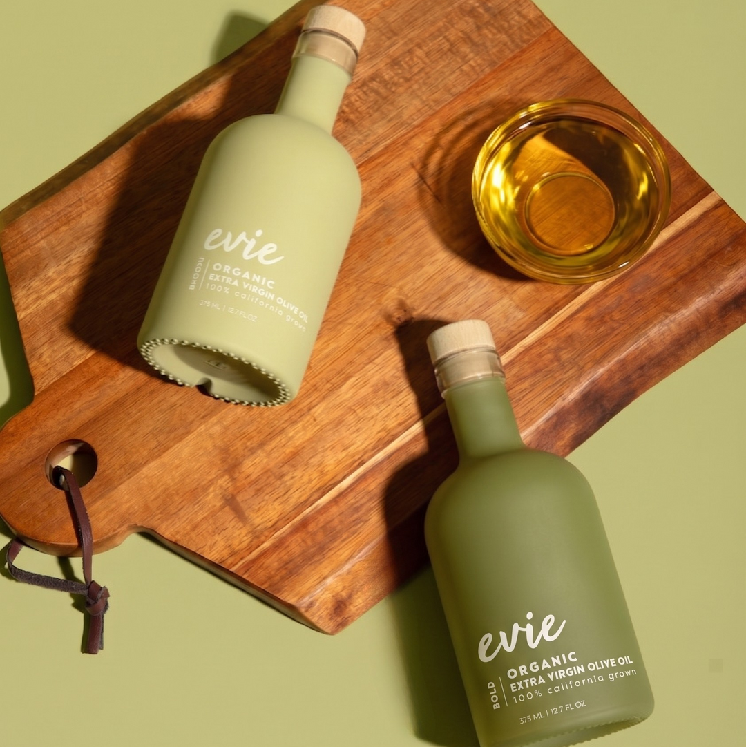 Two green bottles of 'evie' organic olive oil on a wooden cutting board with a glass of olive oil.