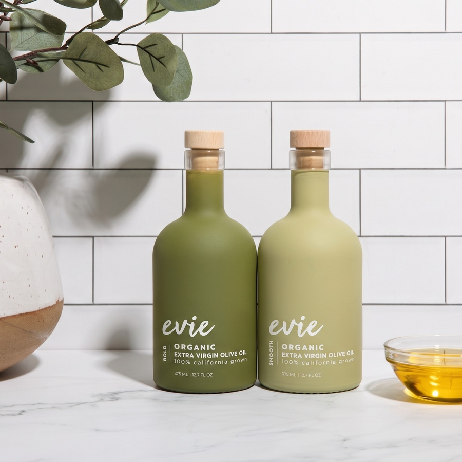 Two bottles of evie organic olive oil on a white surface with a decorative plant and bowl in the background.