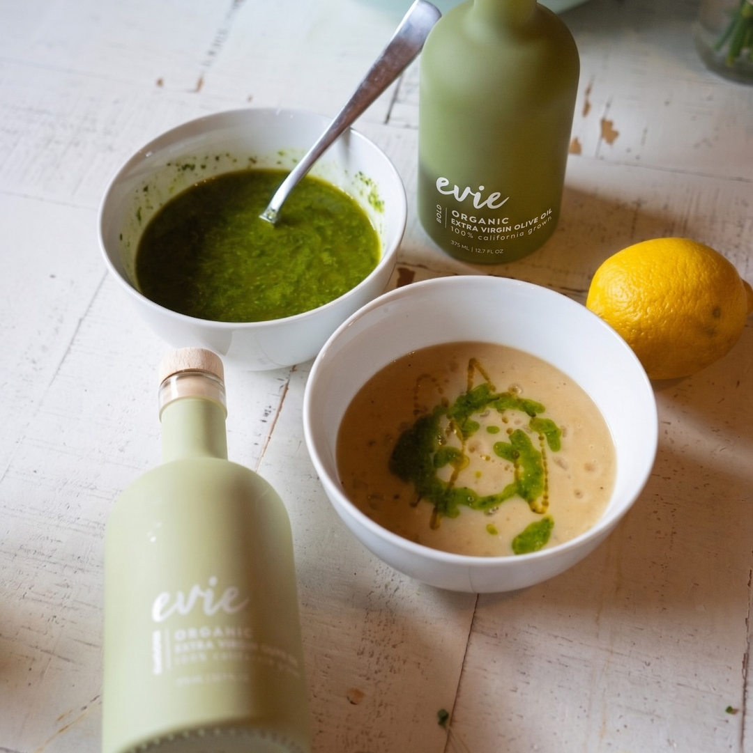 Two bottles of Evie Olive Oil on a wooden surface with bowls of green and brown sauces.