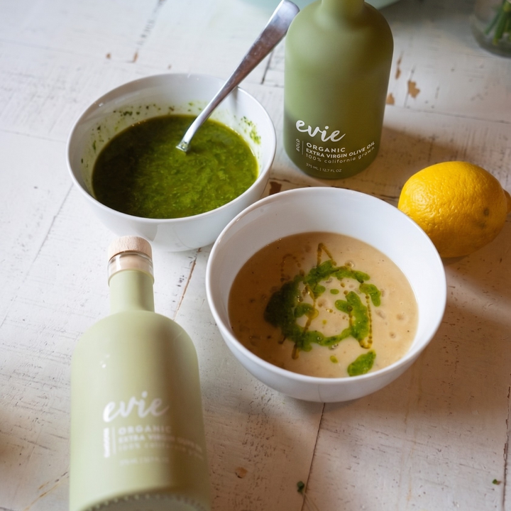 Two bottles of Evie Olive Oil on a wooden surface with bowls of green and brown sauces.